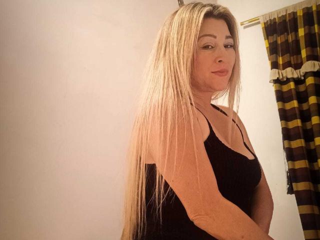 DanaWester with long blonde hair, wearing a black tank top, standing side to camera, looking over shoulder, slight smile, white wall, yellow and brown striped curtain in background.