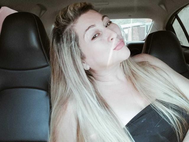DanaWester with long blonde hair, pouty lips, and black strapless top, sitting in a car, head tilted, looking at camera, black leather seats in background.