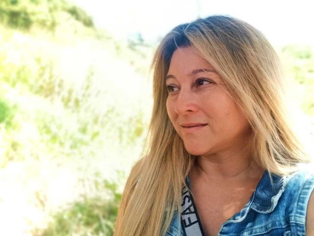 DanaWester with blonde hair, blue denim shirt, smiling slightly, looking to the side, bright sunlight, green blurred background. Her tits are small and her ass is not visible.