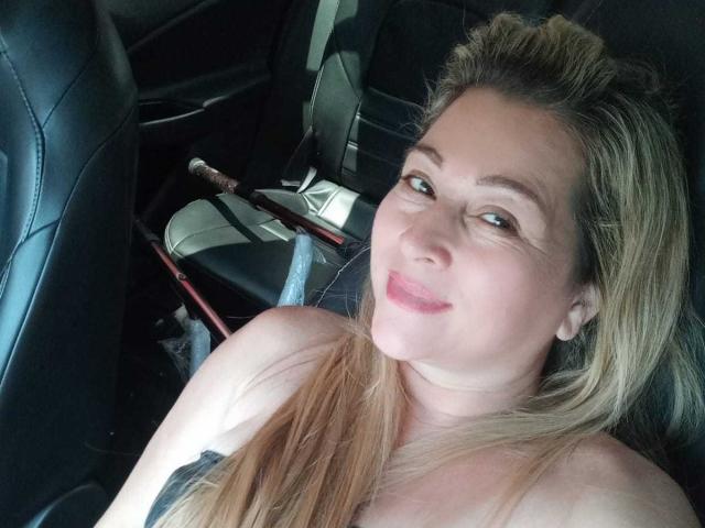 DanaWester with blonde hair, pink lipstick, and a slight smile, sitting in a car, showing her bare shoulders and a black tattoo on her right side.
