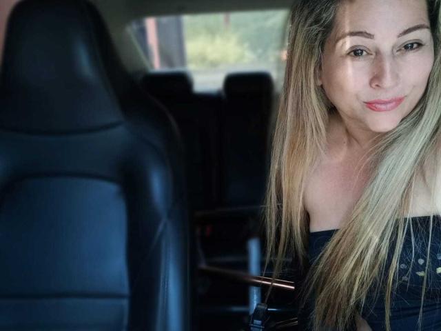 DanaWester has long blonde hair, a cute smile, and is wearing a black off-shoulder top, sitting in a car with a black leather seat in front of her.