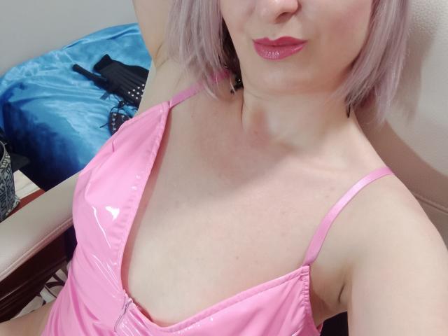 MonicaMiss with pink shiny latex top, showing her tits, pink lips, and pale skin, with pink hair, arm raised, sitting on a chair, blue blanket in background.