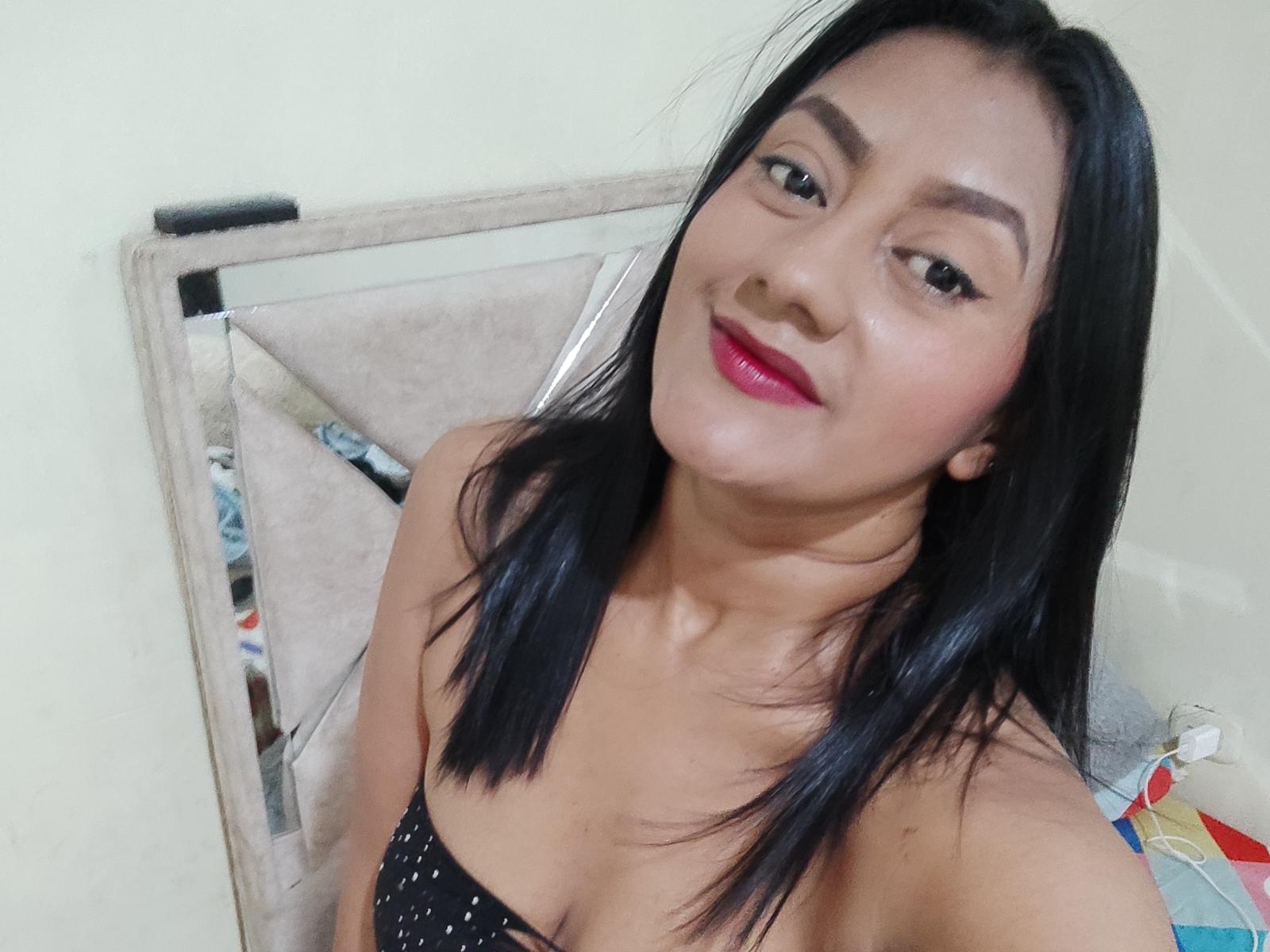 VioletteBlues with black hair, pink lipstick, and a black strapless top showing small tits, smiling at the camera, sitting on a beige chair.