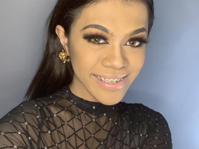 DarkBeautySophia with a smile, wearing a black fishnet top, showing her teeth with braces, brown eyes, long eyelashes, and gold rose earrings. She has tan skin and dark hair.