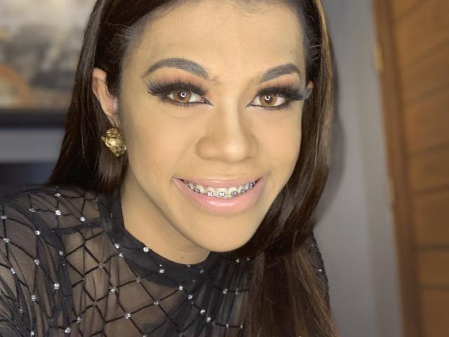 DarkBeautySophia with brown eyes, long brown hair, and a smile showing metal teeth. She's wearing a black sheer top with sparkles, gold earrings, and has heavy eyeliner.