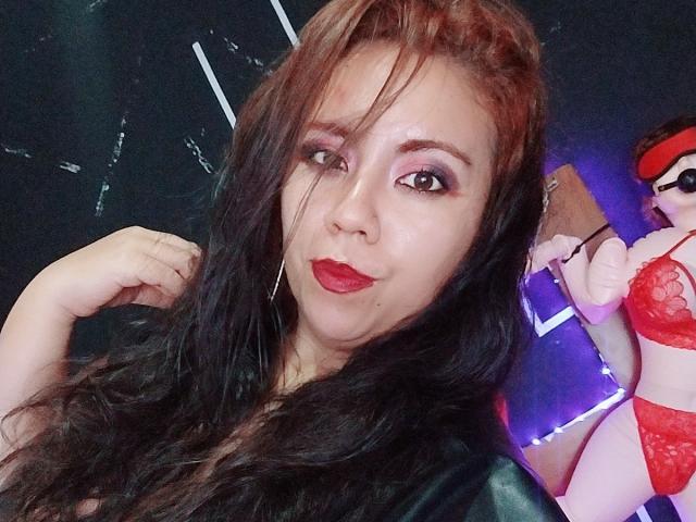LexyDom has long black hair, red lipstick, and pink eye makeup. She's wearing a black leather top, with red lace lingerie in the background. Her hand is near her face.