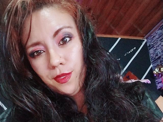 LexyDom has wavy black hair, pink eyeshadow, and red lipstick. She's smiling with a slight pout, showing off her shiny skin and cute nose. Boobs are not visible.