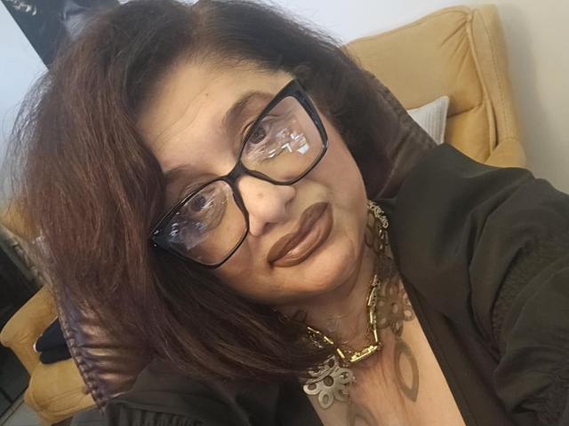 "ClassybutNaughty" mit braunen Haaren, Brille und dunklem Lippenstift, trägt ein schwarzes Oberteil und eine goldene Halskette, lächelt leicht und hat den Kopf leicht geneigt. Ihre Brüste sind teilweise sichtbar..