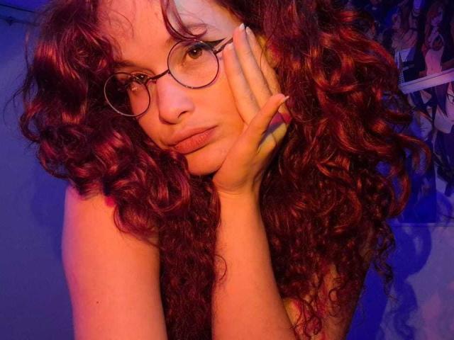 DanaStone with curly red hair, wearing glasses, resting her head on her hand, looking slightly pouty, in a blue-purple lit room, showing her bare shoulder.