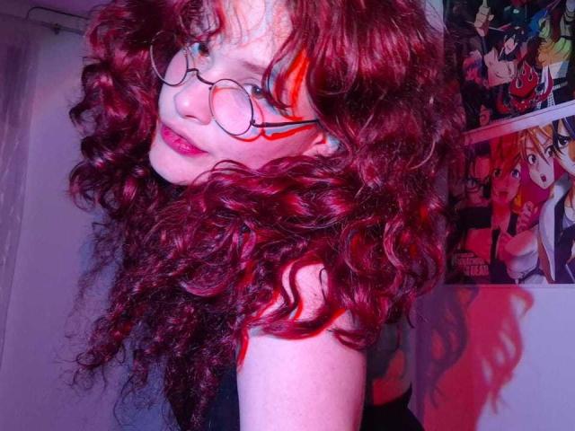 DanaStone with curly red hair, glasses, and pink lipstick, looking over shoulder, showing a cute face and a slight smile, wearing a red top, in a colorful room.