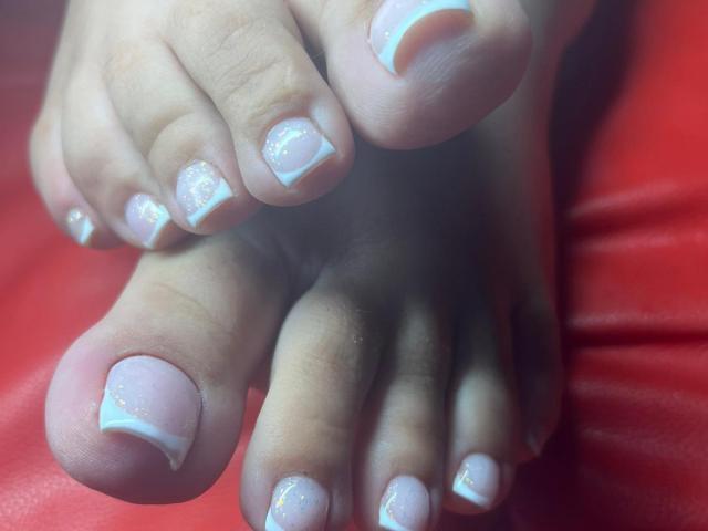 GoddessXDeborah's feet, showing her pinkish toes with white French tips and sparkly nail polish, resting on a red surface. Her toes are slightly curled and close together.