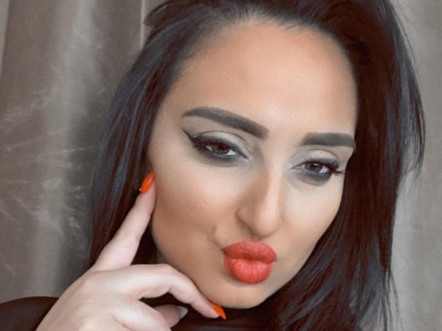 GoddessXDeborah with pouty orange lips, gray eyeshadow, thick eyebrows, long black hair, and orange nails, touching her cheek with a finger. She's looking directly at the camera.