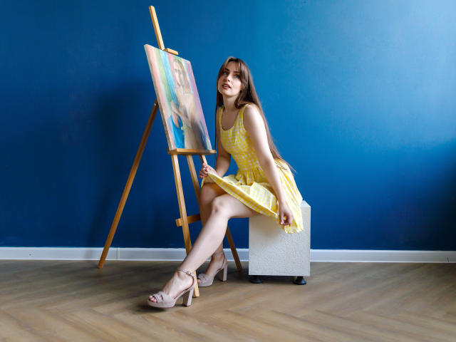 VeronicaTay sits on a stool, wearing a yellow dress, holding a paintbrush, with a canvas on an easel, her legs crossed, showing her ass, in beige heels.