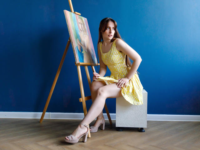 VeronicaTay, with yellow checkered dress and beige heels, sitting on white box, holding painting brush, legs crossed, painting on easel, blue wall, wooden floor, brown hair, serious look.