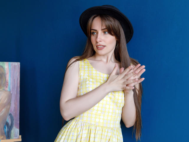 VeronicaTay with long brown hair, wearing a black hat and yellow check dress, standing against a blue wall, hands gently touching her chest, smiling slightly.