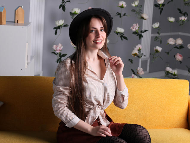VeronicaTay with long brown hair, wearing a black hat, checkered shirt, tied at the front, brown skirt, and black patterned tights, sitting on a yellow sofa, smiling.