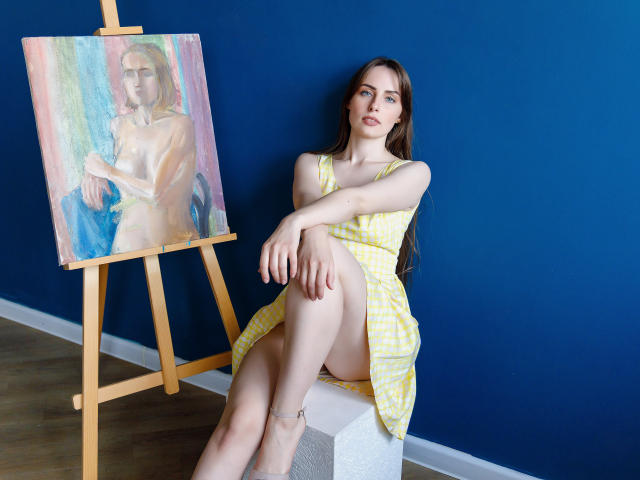 VeronicaTay with long brown hair, wearing a yellow check dress, sitting on a white cube, next to a painting of her nude with small tits and a subtle smile.