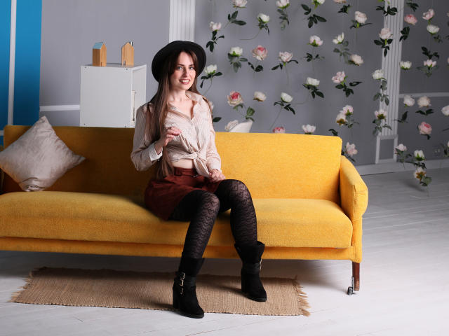 VeronicaTay with tits covered, wearing a black hat, beige blouse, red skirt, black lace tights, black boots, sitting on a yellow couch, smiling.