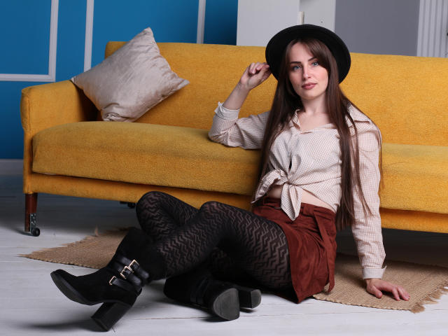 VeronicaTay with long brown hair, black hat, tied beige shirt, brown skirt, black patterned tights, black boots, sitting on floor in front of yellow sofa, hand on hat.