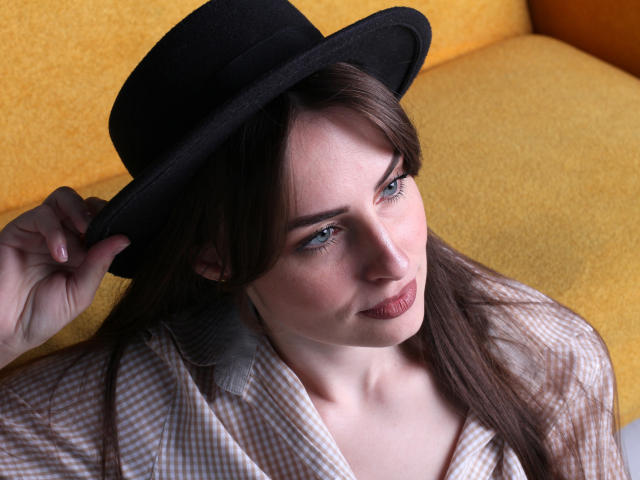 VeronicaTay with blue eyes, wearing a black hat, holding it with her right hand, has long brown hair, light skin, and is dressed in a checkered shirt.