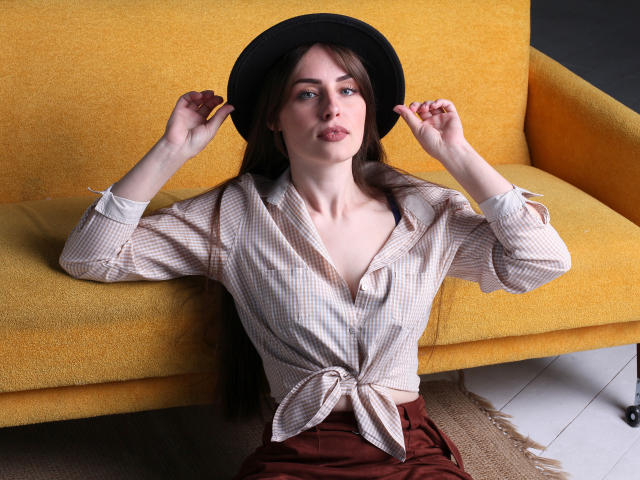 VeronicaTay with long brown hair, wearing a black hat, checkered shirt tied at the front, sitting on a yellow couch, holding her hat with both hands, looking up.