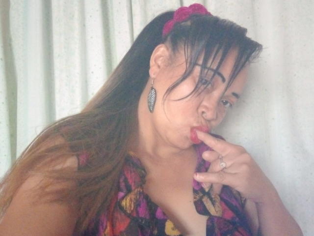 Rahel69, avec des cheveux bruns attachés en queue de cheval, portant un élastique rose, un haut à carreaux et de longues boucles d'oreilles, suce son doigt, avec son doigt dans sa bouche, et montre ses seins..