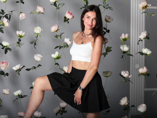 AlenaBlare has tits in a white crop top, black skirt, and a necklace, with one leg raised, smiling at the camera, wearing a black watch.