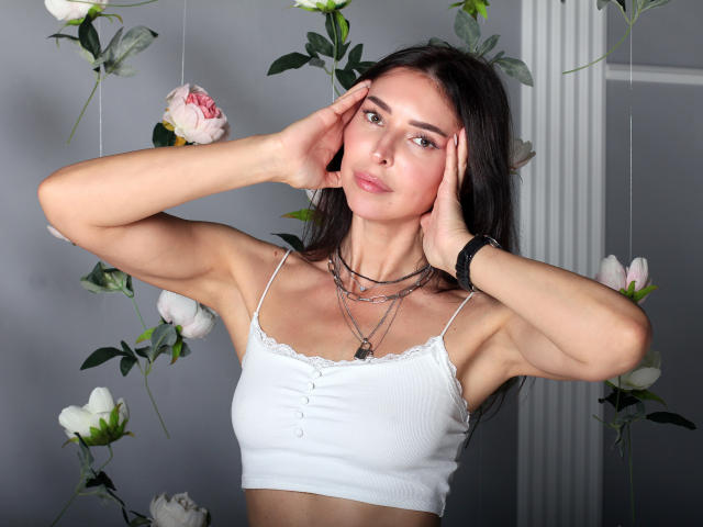 AlenaBlare with hands on her face, wearing a white spaghetti-strap top with lace trim, showing her tits, brown hair, and a black wristband, surrounded by hanging flowers.