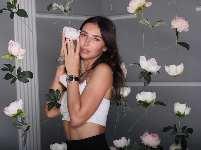 AlenaBlare with long brown hair, white crop top, black watch, holding pink peonies, soft expression, gray background, surrounded by hanging flowers.