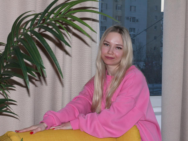 JuliaMacey with blonde hair, pink hoodie, and red nails, sitting on yellow couch, leaning on plant, smiling softly. Her pink tits are slightly visible under hoodie.