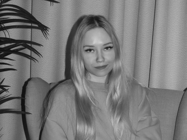 JuliaMacey with long blonde hair, wearing a loose shirt, sitting in a chair, looking slightly to the side with a subtle smile, and a plant on the left.