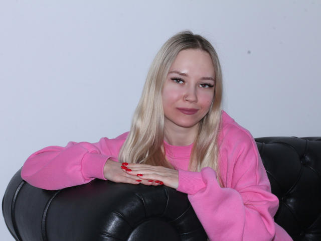 JuliaMacey with blonde hair, pink sweatshirt, red nails, leaning on black leather couch, smirking slightly, medium tits, fair skin, white background, relaxed pose, casual style.