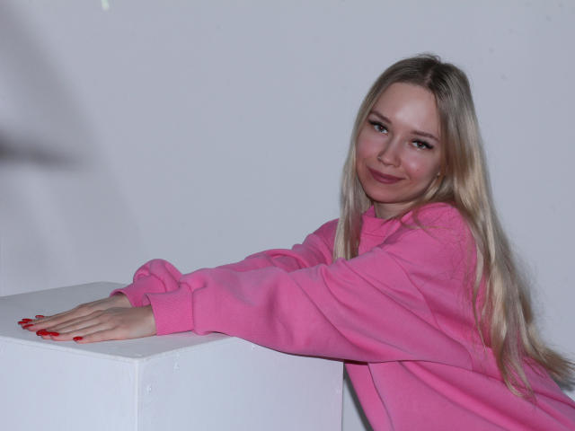 JuliaMacey with long blonde hair, pink jacket, red nails, and a slight smile, leaning on a white cube, showing her cute face and subtle ass.