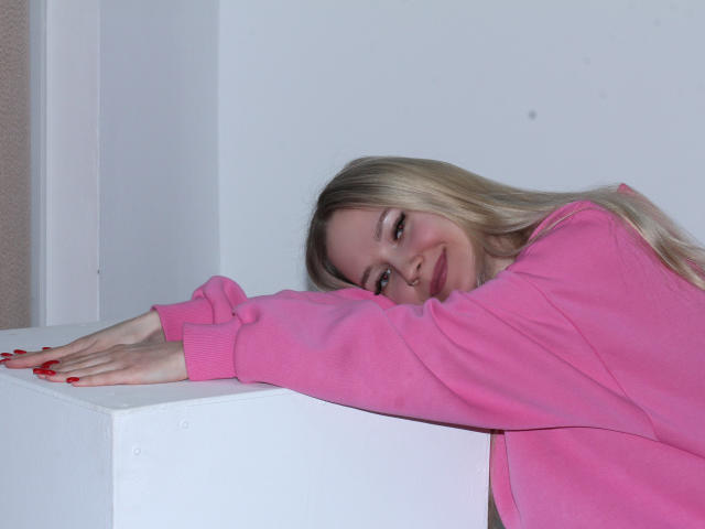 JuliaMacey with long blonde hair, pink sweater, red nails, leaning on white cube, smiling softly, eyes half-open, right arm resting on cube, looking relaxed and cute.