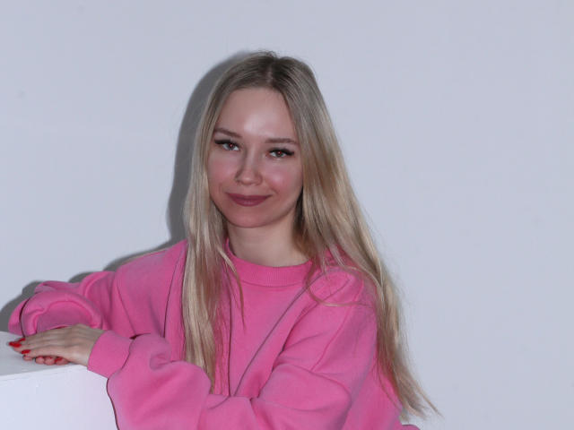 JuliaMacey with long blonde hair, pink sweater, and red nails, smiling softly. She's leaning on a white surface, showing her cute face and pink lips.