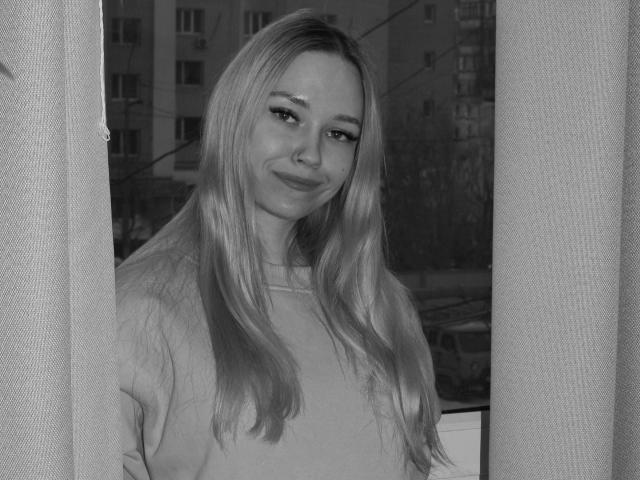 black-and-white photo of JuliaMacey with long blonde hair, smiling, wearing a loose shirt, standing in front of a window with curtains, showing a cute, relaxed face.