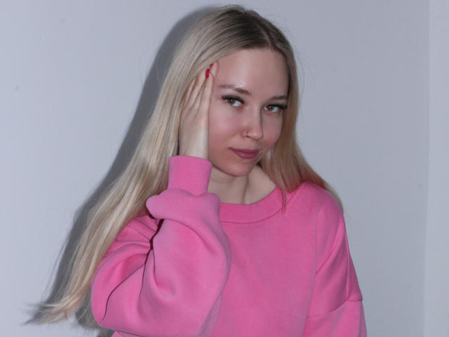 JuliaMacey with long blonde hair, pink sweater, red nail polish, hand on cheek, slight smile, soft makeup, white background, light skin, subtle shadow.