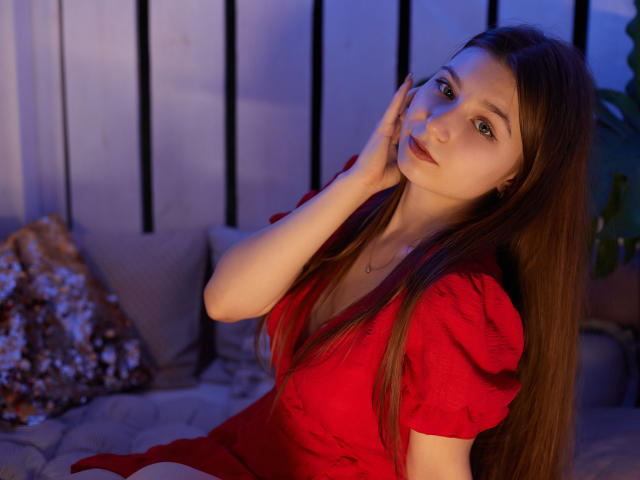 Rina Lewin, con i suoi lunghi capelli castani, il vestito rosso e le labbra piene, che si tocca il viso, seduta su un letto con cuscini scintillanti, illuminata da una luce soffusa blu e rossa..