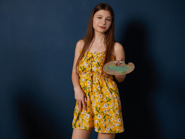 RinaLewin with long brown hair, yellow floral dress, holding a paint palette, smiling, standing against a dark blue wall, wearing a necklace.