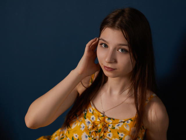 RinaLewin with long brown hair, green eyes, and a yellow floral dress, touching her ear, wearing a delicate necklace, looking at the camera with a slight smile.