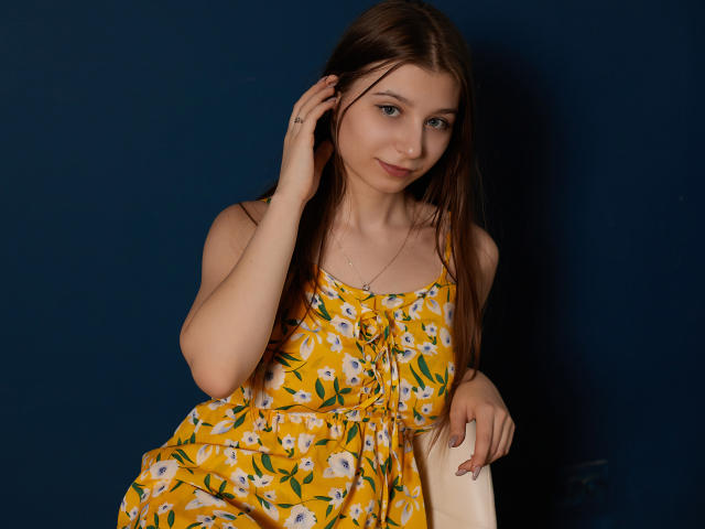RinaLewin with long brown hair, yellow floral dress, small tits, blue eyes, slight smile, hand in hair, sitting, dark blue background, white bag in her right hand.