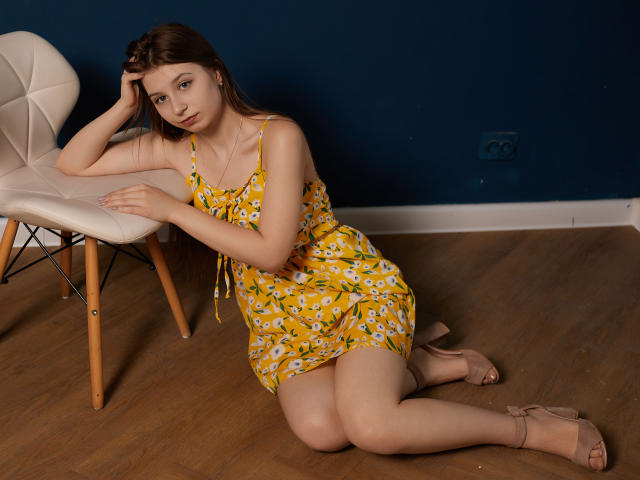 RinaLewin mit kleinen Brüsten, sitzend auf einem Holzboden, trägt ein gelbes Blumenkleid und beige High Heels, lehnt sich an einen Stuhl, blaue Wand, braunes Haar, blickt in die Kamera, Hand auf dem Kopf..