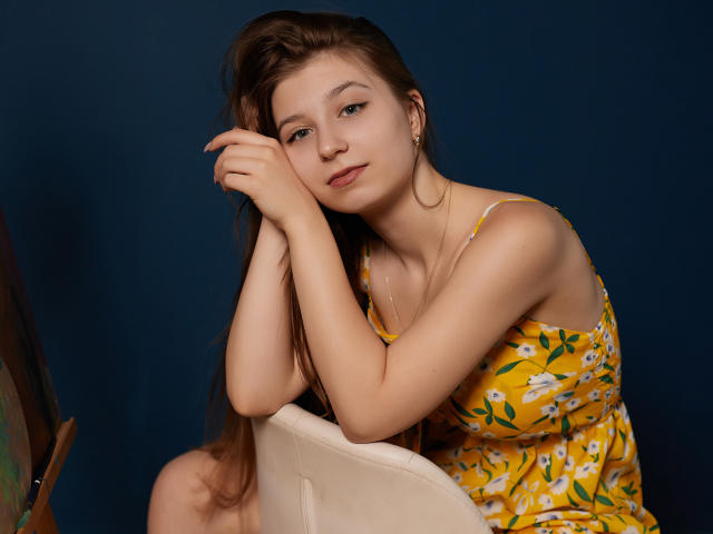 RinaLewin with long brown hair, wearing a yellow floral dress, leaning on a white chair, arms crossed, subtle smile, dark blue background, looking slightly to the right.