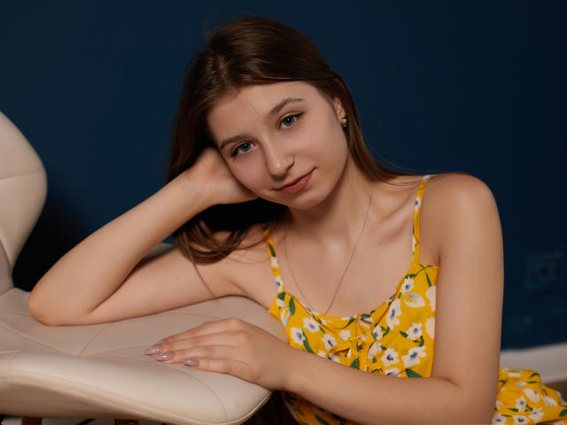 Rina Lewin, con la pelle chiara e i capelli castani, indossa un abito giallo con fiori, è appoggiata a una sedia bianca, sorride leggermente, con una mano sul viso, le unghie dipinte, sfondo blu..
