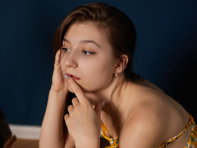 RinaLewin tiene la piel clara, el pelo castaño y los ojos azules, lleva una blusa con estampado floral, y tiene un dedo sobre su rostro ligeramente entreabierto, con labios brillantes y una expresión adorable, sobre un fondo azul oscuro..