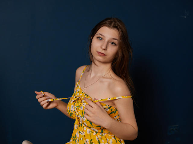 Rina Lewin, con i capelli castani, indossa un abito floreale giallo, sta tirando giù le bretelle, mostrando piccoli seni, ha la pelle chiara, un sorriso appena accennato, sullo sfondo c'è un colore blu scuro, è in piedi, in posa casual, leggermente sexy, con una luce soffusa..