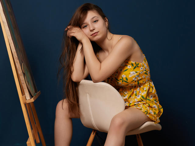 RinaLewin with long brown hair, sitting on a white chair, wearing a yellow floral dress, arms resting on her head, looking slightly bored, against a dark blue background.