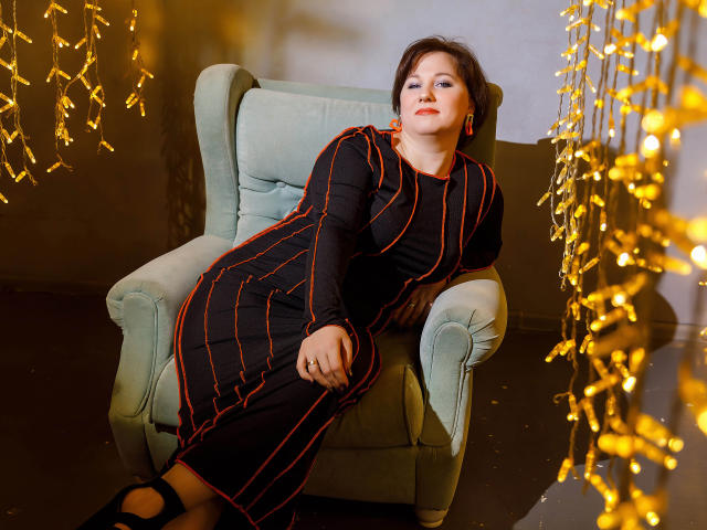 MirandaBrooks is sitting in a green armchair, wearing a black dress with orange stripes, looking relaxed with a slight smile. Golden string lights hang in the foreground.