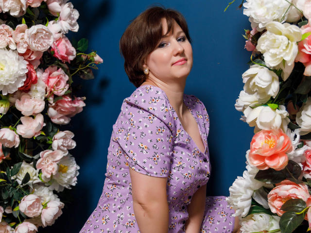 MirandaBrooks with short brown hair, wearing a lilac dress with white daisy patterns, sitting between floral arrangements, pink and white roses, blue background, looking up with a slight smile.