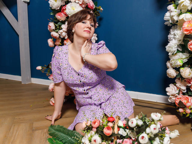MirandaBrooks, with short brown hair, wears a purple floral dress, kneeling on wooden floor, surrounded by roses, touching her neck, smiling, in a blue-walled room.